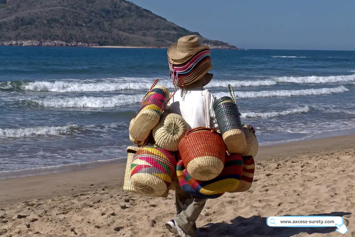 A man selling straw hats and baskets carries his heavy load along a sandy beach.