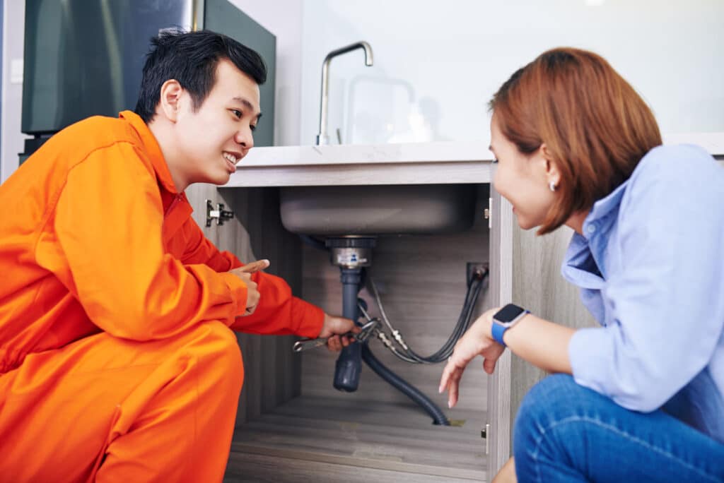 Smiling Vietnamese plumber showing fixed or installed pipes under sink in kitchen of young woman