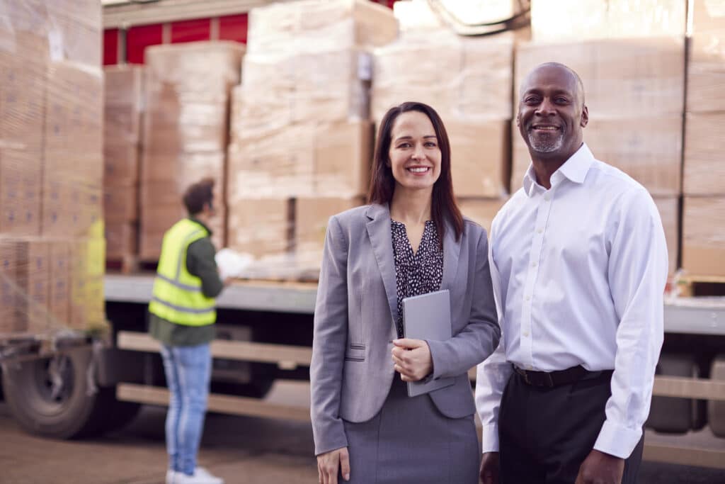Portrait Of Multi-Cultural Freight Haulage Team Standing By Truck Being Loaded By Fork Lift