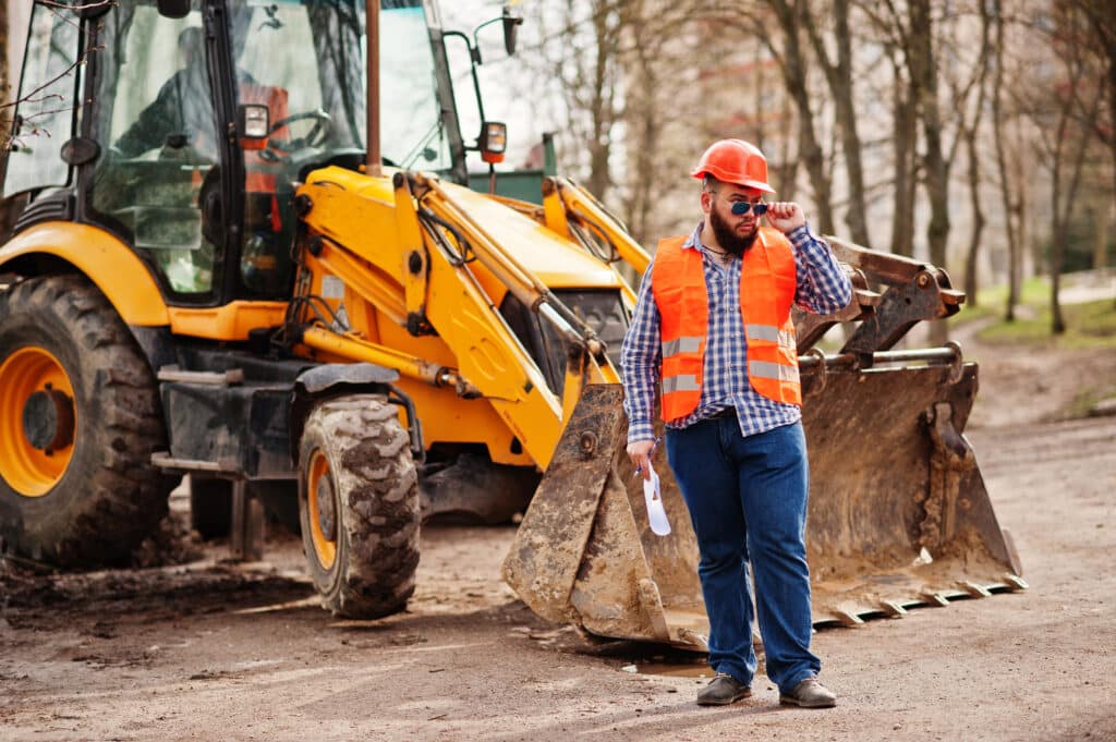 Brutal beard worker man suit construction worker in safety orange helmet, sunglasses against traktor with plan paper at hands.