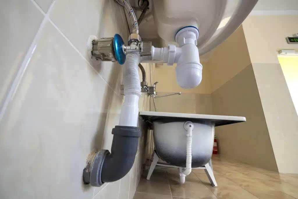 Bottom view of new white washbasin sink connected to sewer on background of bathroom light beige ceramic tiles. Professional plumbing and installation concept.