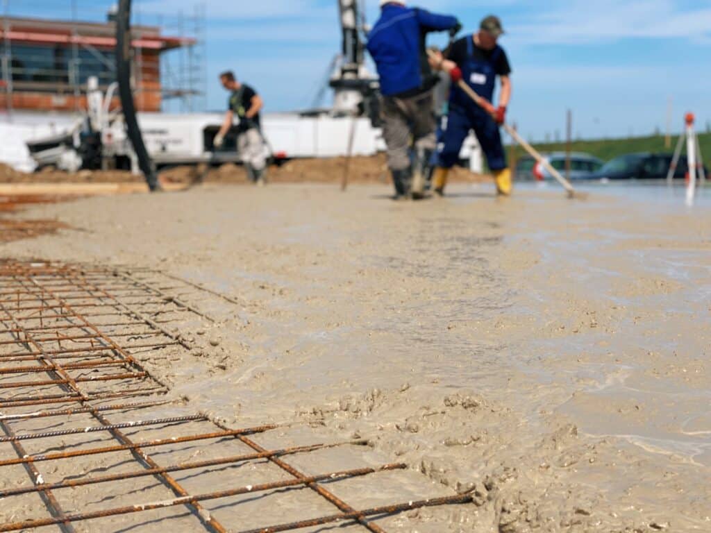 Full length of man laborer using troweling machine while screeding floor in new building under blue sky. Male worker finishing concrete surface with floor screed grinder machine at construction site.