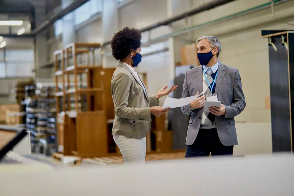 Mid adult businessman communicating with African American colleague while visiting production line in a wood factory during COVID-19 pandemic.