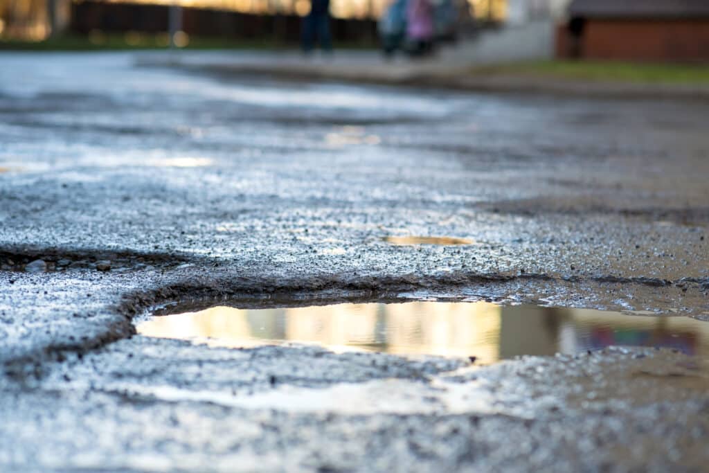 Close up of a road in very bad condition with big potholes full of dirty rain water pools.