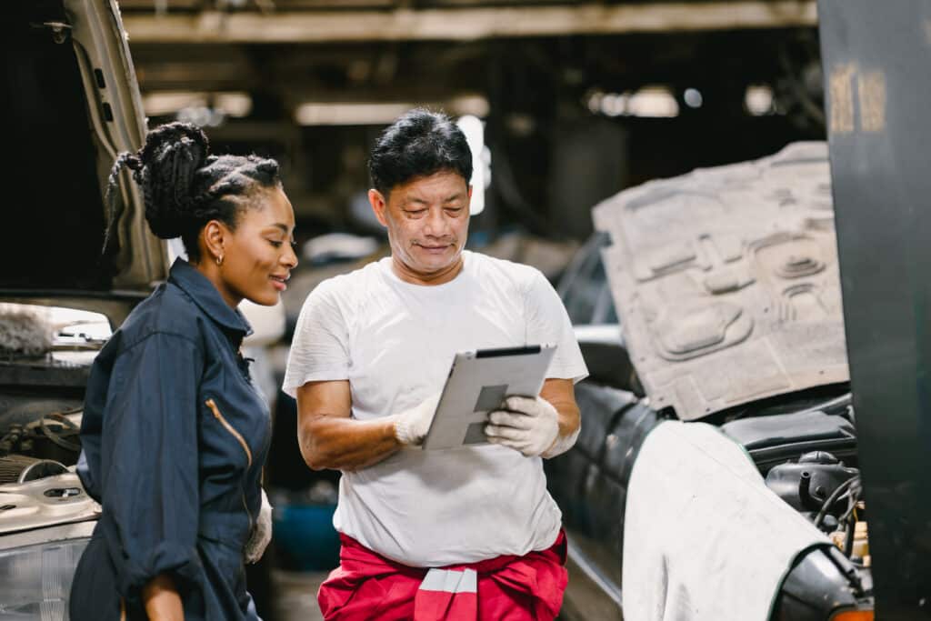 Mature expert mechanic man training young teen women worker to check service car using tablet technology.