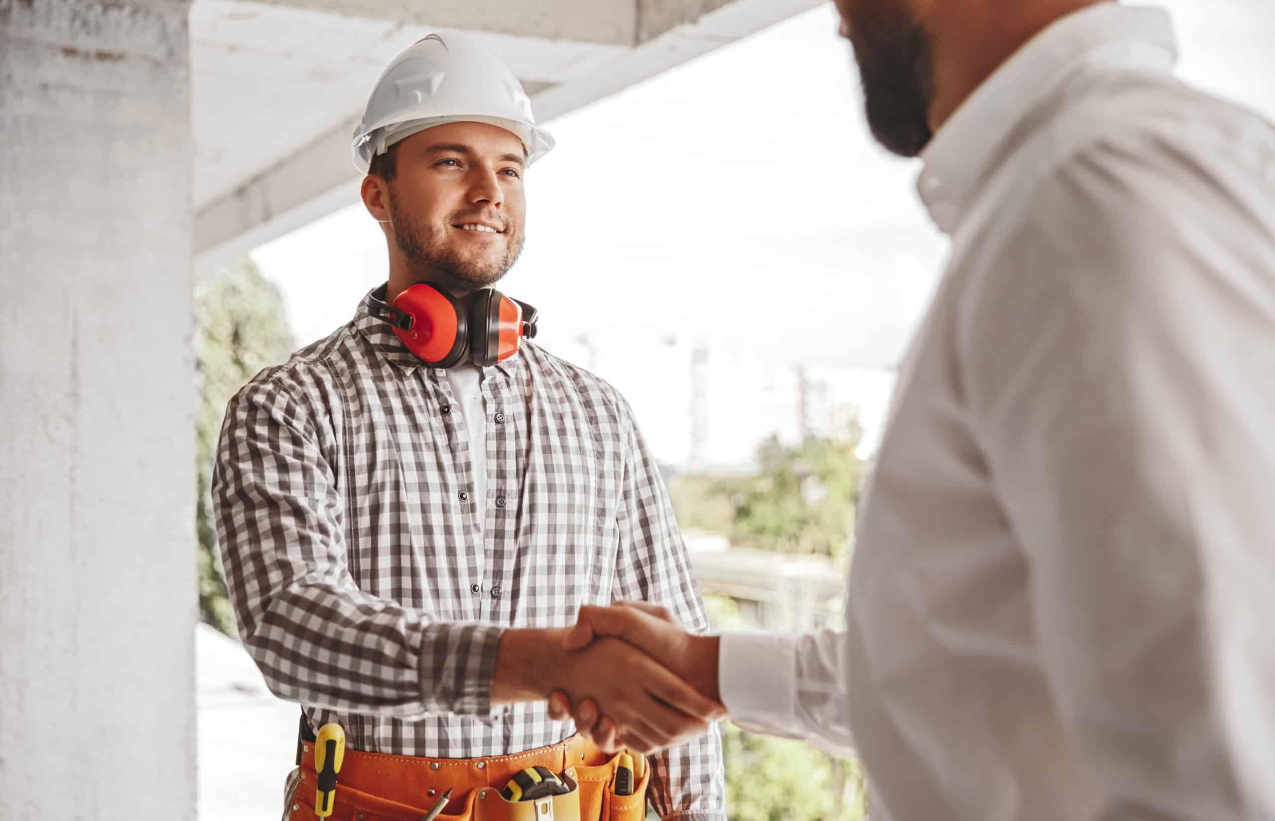 Positive young male engineer in protective hardhat shaking hand of partner while greeting each other on construction site
