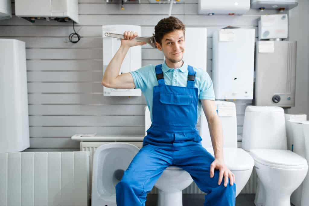 Plumber with pipe wrenches sitting on toilet at the showcase in plumbering store. Man buying sanitary engineering tools and equipment in shop