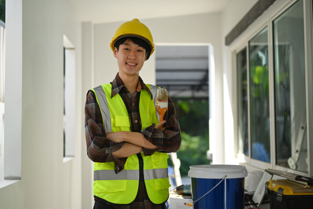 Professional painter in safety helmet holding painting brush standing in construction site and smiling at camera.