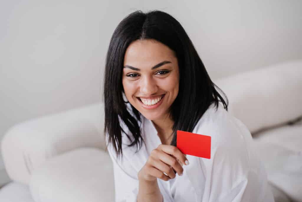 Cheerful Young African american entrepreneur with straight hair and toothy smile holding credit card, satisfied by her growing business. Happy successful girl sitting on sofa at home. Money concept.