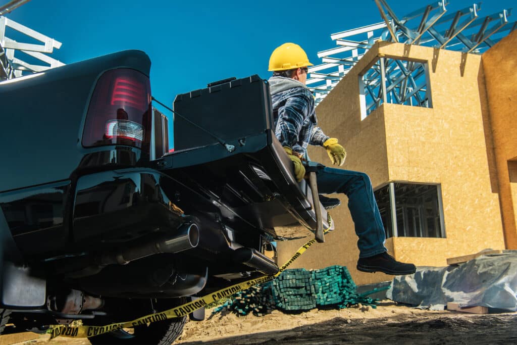 Tired Contractor Sitting on His Pickup Truck Cargo Bed Taking a Moment of Rest During Hard Day at Work. Construction Site in the Background. Industrial Theme.