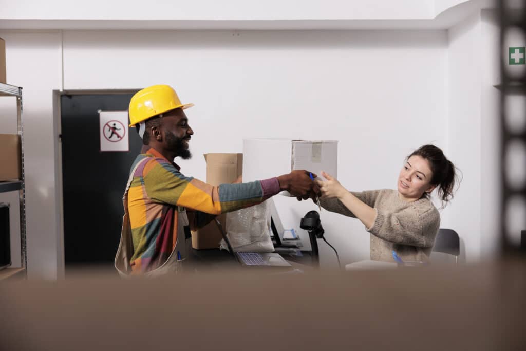 Diverse stockroom team working at customers orders, preparing packages for delivery in storage room. African american wearing industrial overall and helmet working at goods inventory