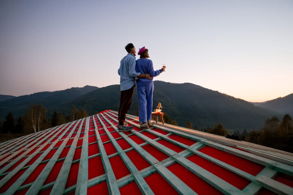 Man and woman enjoy beautiful sunset in the mountains, while standing on the roof of their unfinished house. Concept of romance, house of dream and escaping to nature