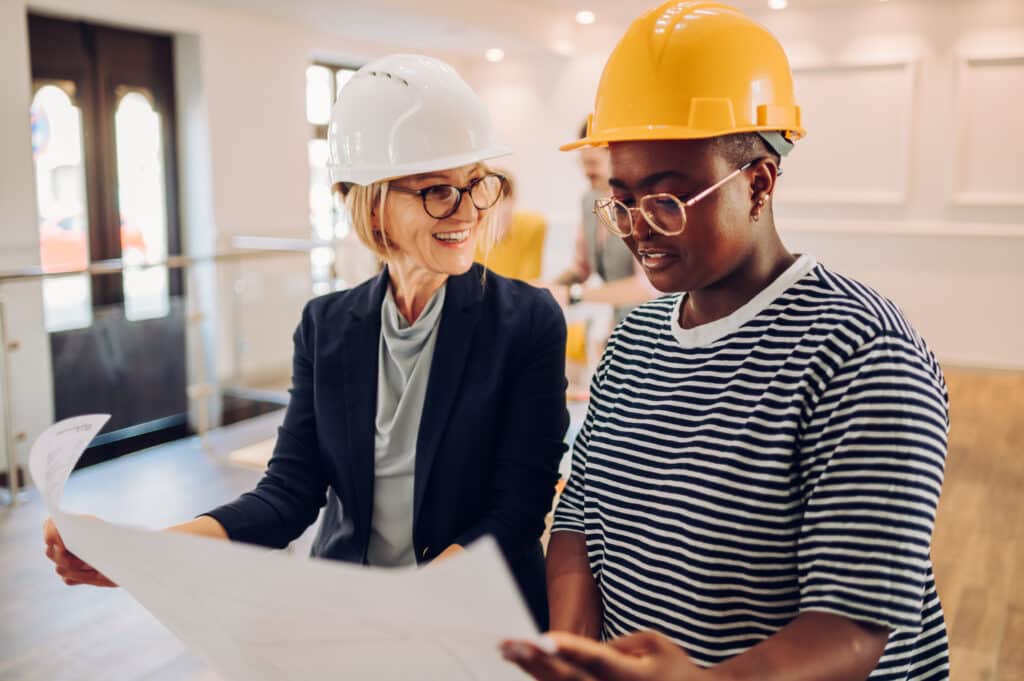 Team of two architects working on house plan project in an office while wearing helmets and holding blueprints. Senior female boss and african american woman holding rolled up building plans.