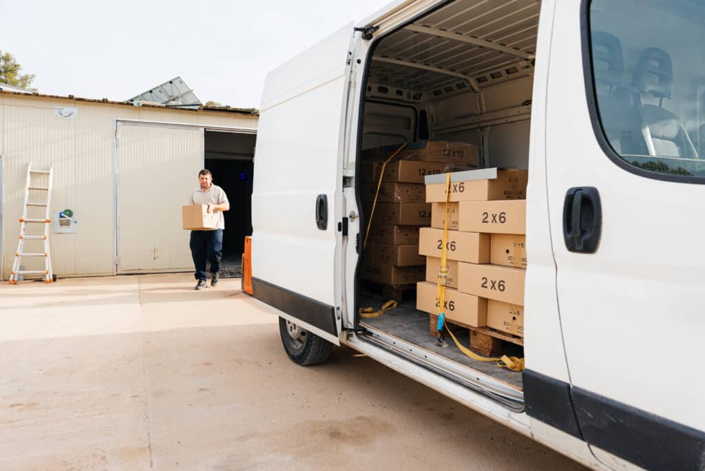 Concentrated young male walking with carton box in hands near warehouse walking towards parked van with open door and loaded cardboard boxes