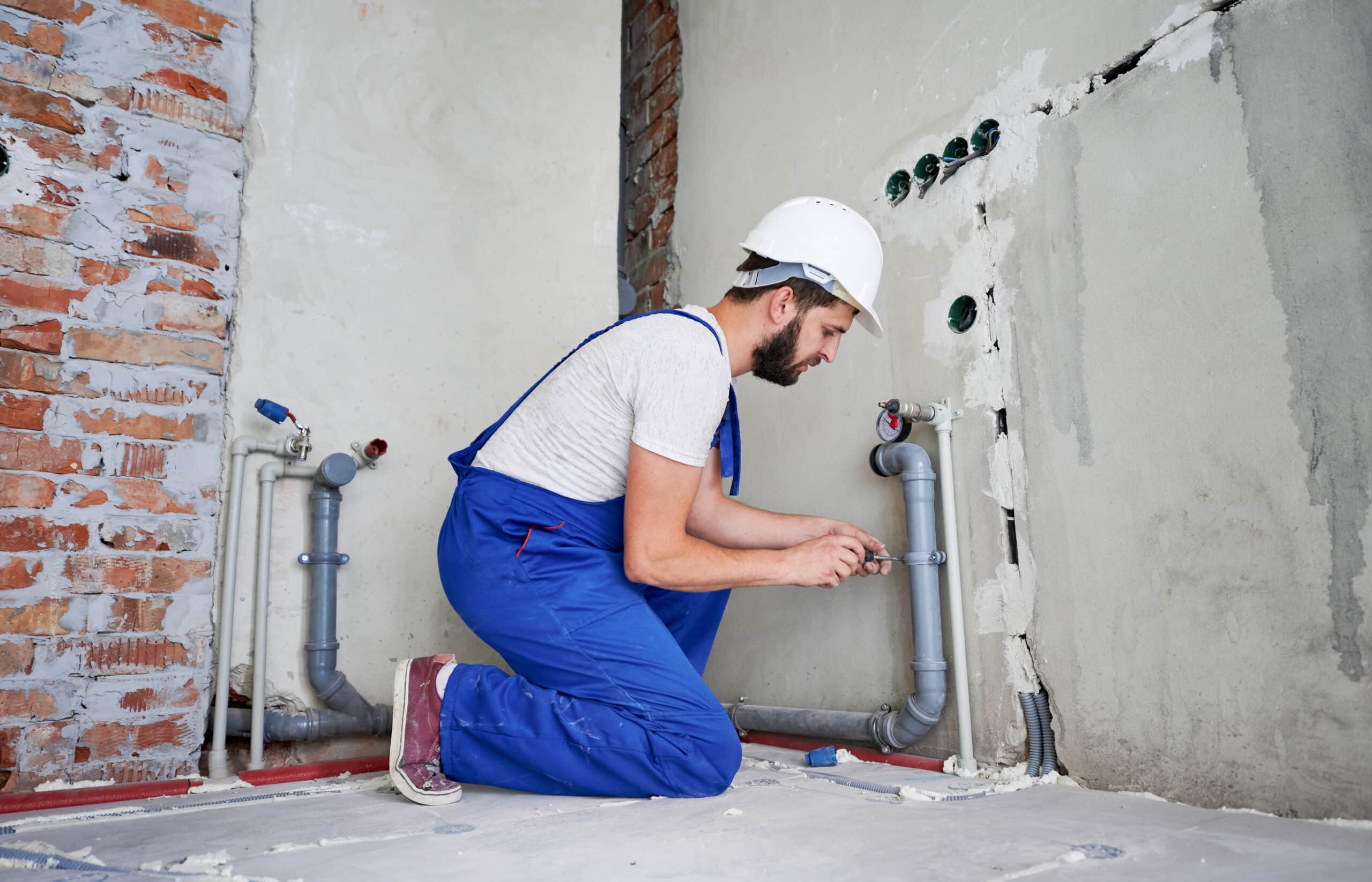 Horizontal snapshot of young plumber working with grey sewer pipes, fixing them to wall with a help of screwdriver. Side view of plumber standing on knees wearing blue uniform and white helmet