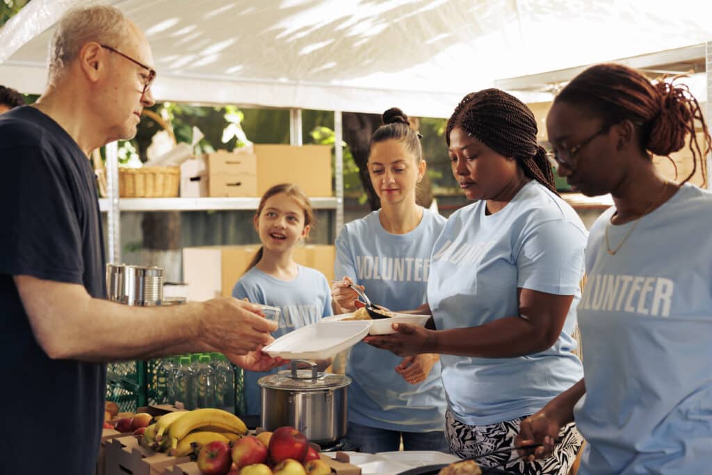 Multiracial volunteer women at local center giving food assistance serving meals and non-perishable items to hungry and homeless people. Friendly charity workers feed warm meals to underprivileged.