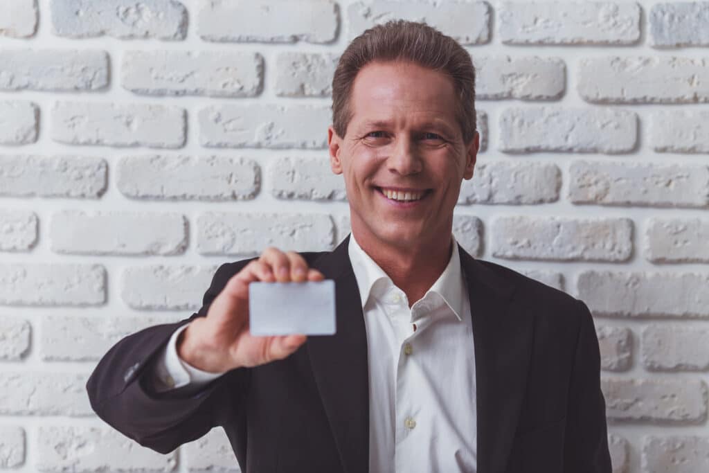 Handsome middle aged businessman in classic suit is holding business card and smiling, standing against white brick wall