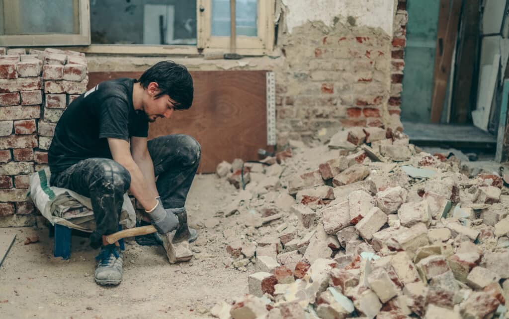 A young Caucasian brunette man with a beard in black work clothes sits on a small chair on the left and cleans the bricks from dirt, debris and old cement with an ax, close-up side view.Construction work concept.