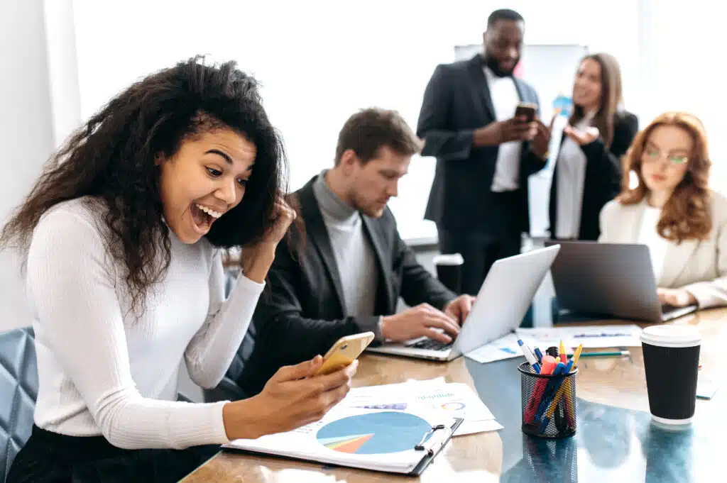 Overjoyed female employee is sitting in office, looking at smartphone screen, smiling. Excited african american business woman well done with project or startup, receive big profit, success concept