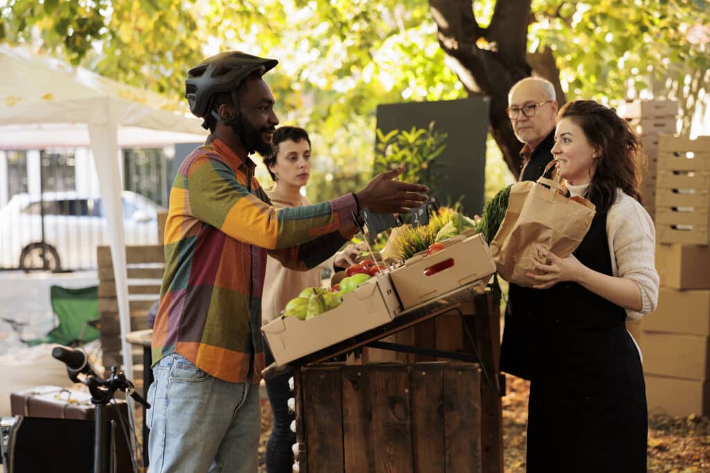 Farmers market vendors providing organic food delivery, deliveryman helping with produce shipping. Family of local farmers standing behind fruits and vegetables stand at food market.
