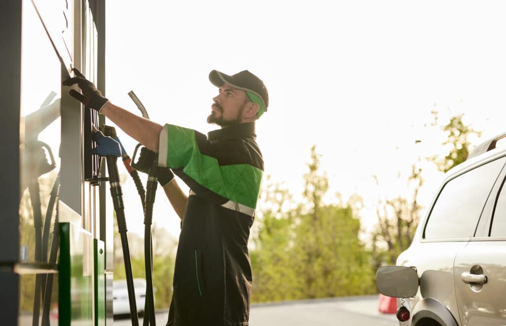 Side view of concentrated young bearded male employee in uniform pushing buttons and holding gas pump nozzle before refueling modern SUV car during work at filling station