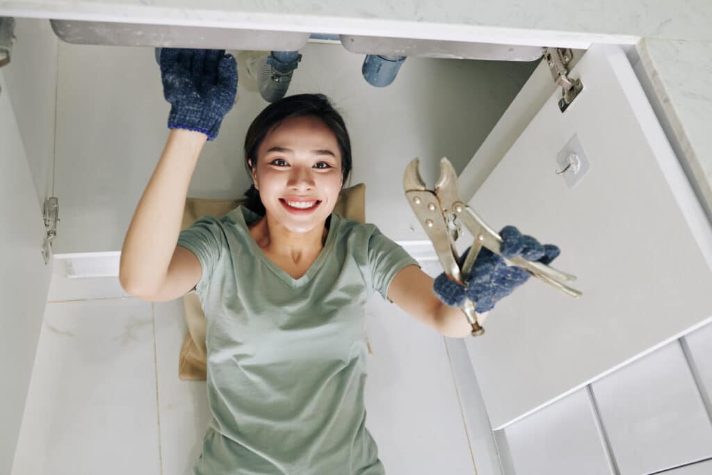 Smiling young pretty Vietnamese woman using wrench when fixing pipe problem in her kitchen