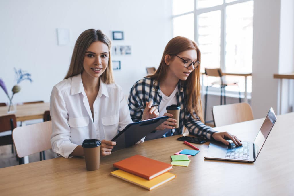 Positive young businesswoman freelancer sitting with female coworker with coffee cups at table with laptop and looking at screen while working on project and colleague looking at camera with clipboard in hand