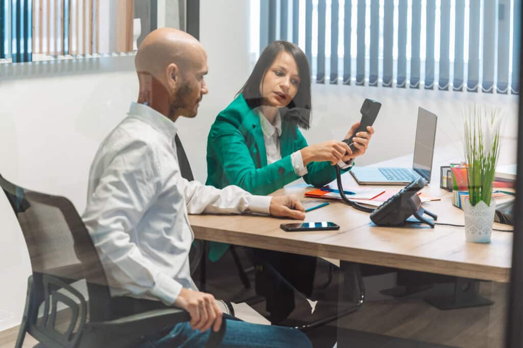 A business meeting in a modern office setting. A bald man and a woman in a green blazer are discussi