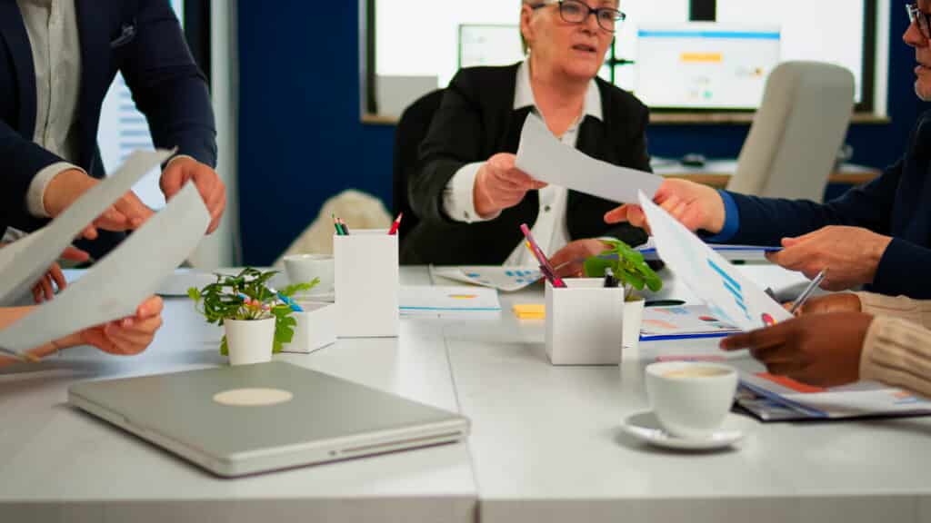 Confident company manager giving working tasks to diverse teamworkers analysing paperwork with graphs sitting in start up office. Multiethnic team discussing project ideas at brainstorming meeting