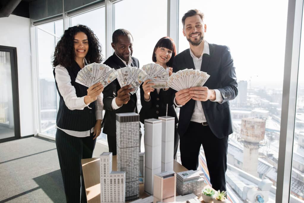 Team of diverse multiethnic business people holding bundle of american dollar cash money, staying near table with 3d skyscraper model at office interior with panoramic city view.