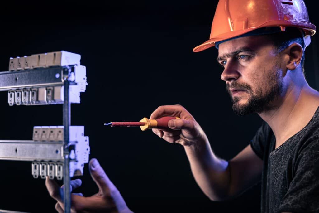 A male electrician in a protective helmet works in a switchboard with an electrical connecting cable.