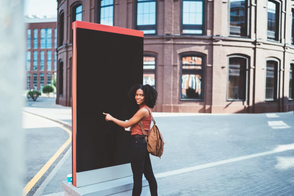 Back view of black cheerful traveler with Afro hairstyle in casual clothes with bag pointing at billboard exploring city on sunny day