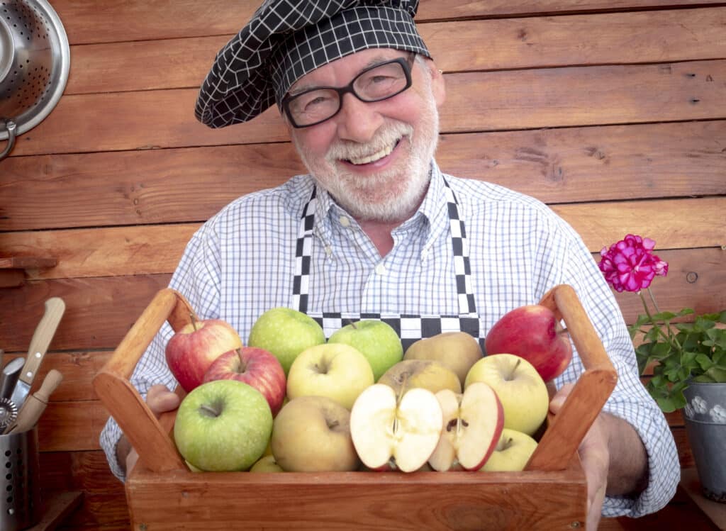 Portrait of smiling senior man with black eyeglasses holding a basket full of apples of various qualities and colors. One retired people. Rustic background in recycled wood. Healthy eating concept
