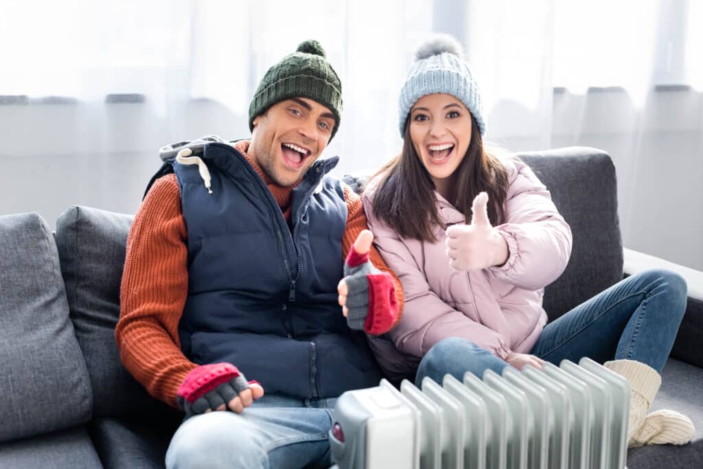 smiling girlfriend and boyfriend in winter outfit showing thumbs up and warming up near heater