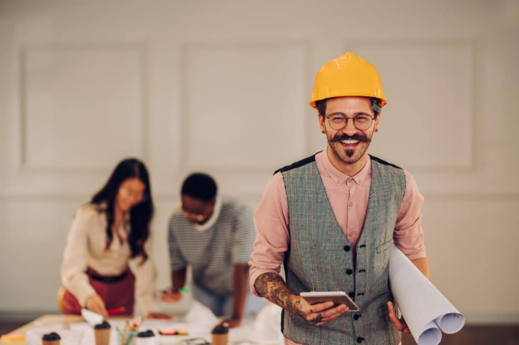 Portrait of smiling man architect with blueprints and a tablet in creative office. Tattooed male contractor holding roll of architectural projects and looking into the camera. Copy space.